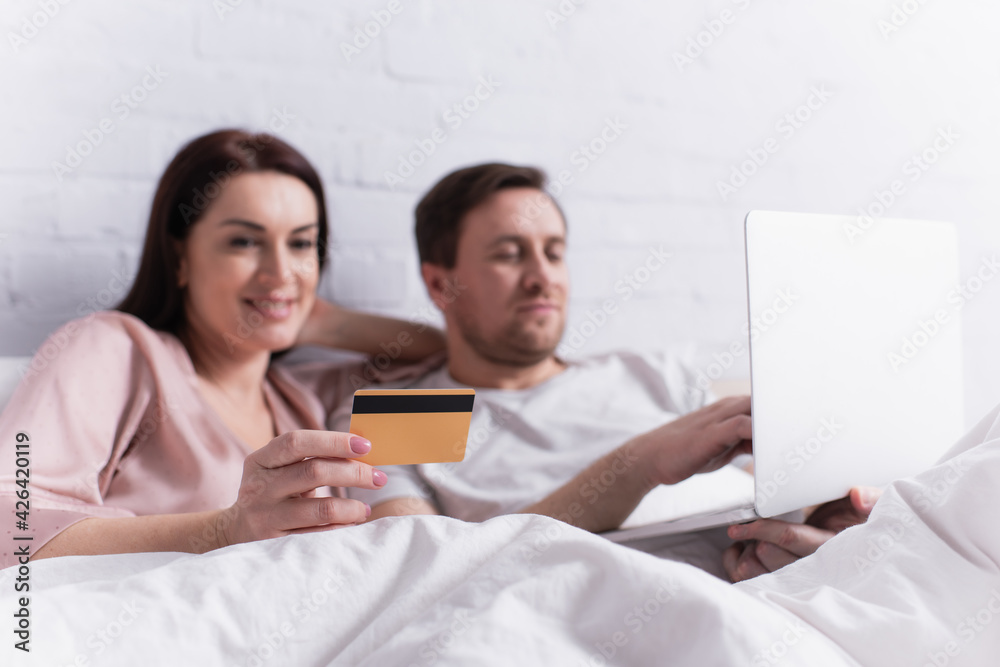 Fototapeta premium Credit card in hand of woman lying near husband with laptop on blurred background on bed