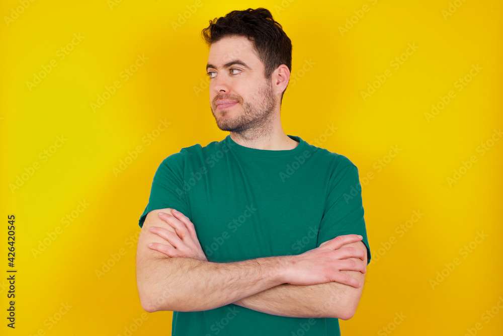 Pleased young handsome caucasian man wearing green t-shirt against yellow background keeps hands crossed over chest looks happily aside Pleased young handsome caucasian man wearing green t-shirt against yellow background keeps hands crossed over chest looks happily aside