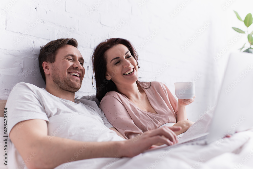 Smiling man using laptop on blurred foreground near wife with cup on bed Smiling man using laptop on blurred foreground near wife with cup on bed