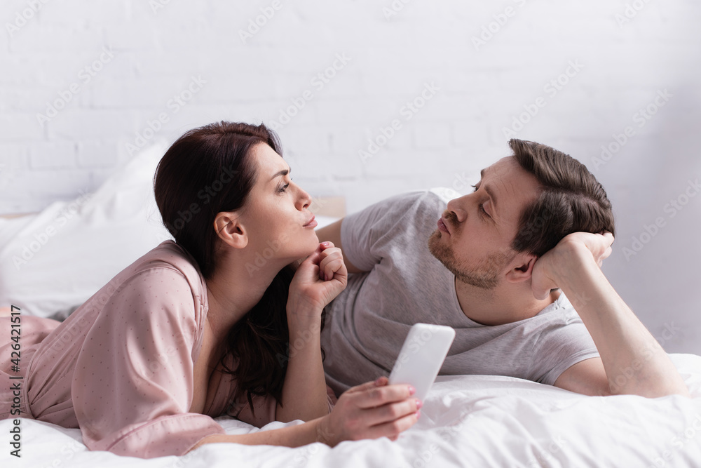 Woman with smartphone pouting lips near husband on bed Woman with smartphone pouting lips near husband on bed