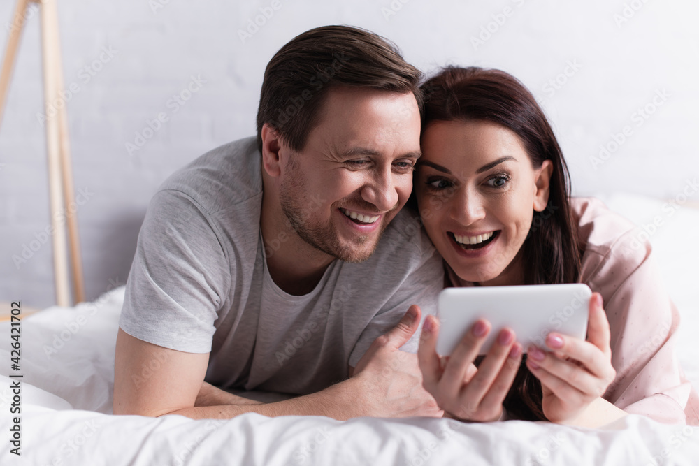 Smiling woman holding cellphone on blurred foreground near husband in bedroom Smiling woman holding cellphone on blurred foreground near husband in bedroom