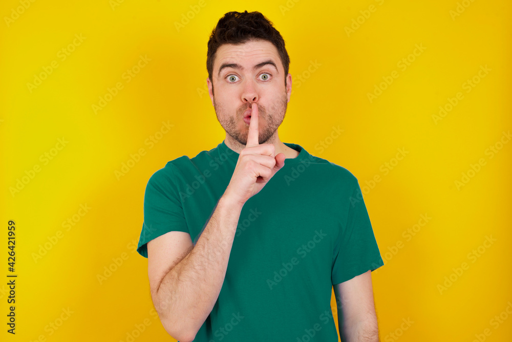 Surprised young handsome caucasian man wearing green t-shirt against yellow background makes silence gesture, keeps finger over lips and looks mysterious at camera Surprised young handsome caucasian man wearing green t-shirt against yellow background makes silence gesture, keeps finger over lips and looks mysterious at camera