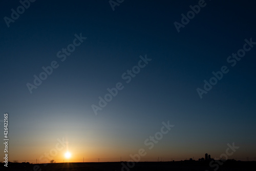 A setting sun silhouettes the horizon, distant farm buildings and silos.