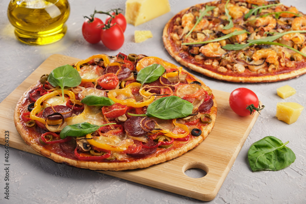 Two homemade pizzas with mozzarella cheese, tomatoes, mushrooms, salami and seafood decorated with basil and arugula on a gray background. Close-up, horizontal orientation without people. Two homemade pizzas with mozzarella cheese, tomatoes, mushrooms, salami and seafood decorated with basil and arugula on a gray background. Close-up, horizontal orientation without people.