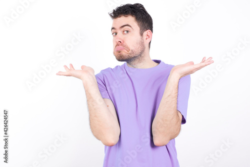 Puzzled and clueless young handsome caucasian man wearing purple t-shirt against white background with arms out, shrugging shoulders, saying: who cares, so what, I don't know.