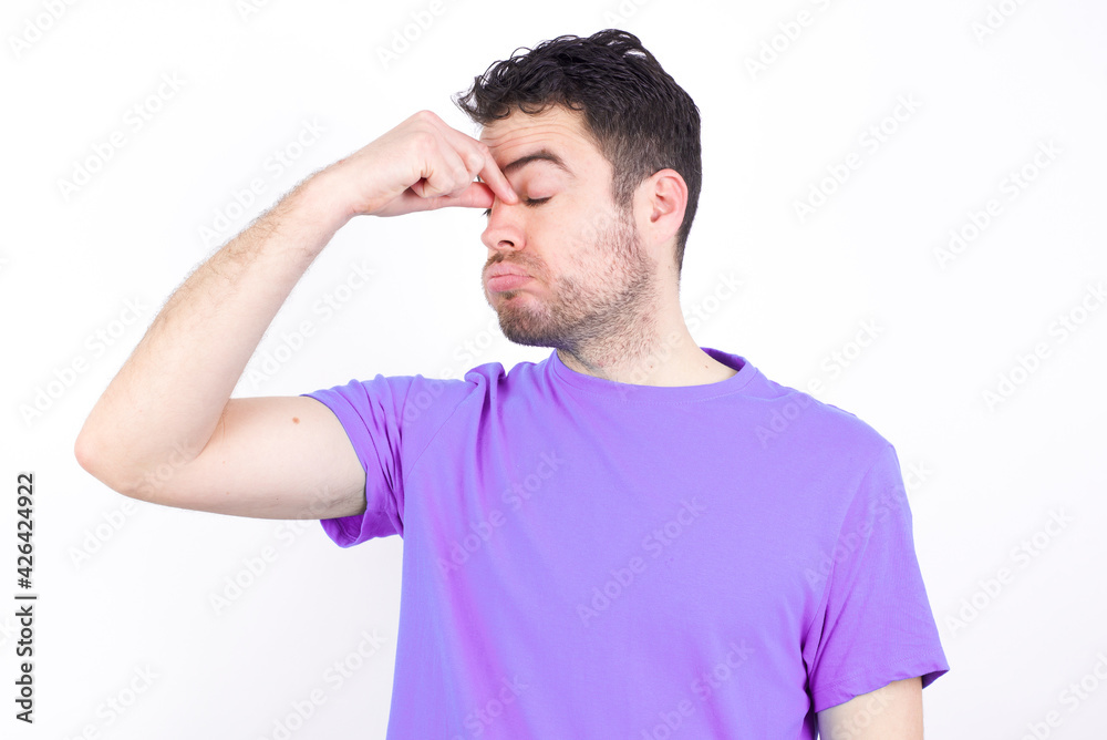 Very upset, young handsome caucasian man wearing purple t-shirt against white background touching nose between closed eyes, wants to cry, having stressful relationship or having troubles with work Very upset, young handsome caucasian man wearing purple t-shirt against white background touching nose between closed eyes, wants to cry, having stressful relationship or having troubles with work