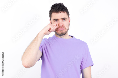 Unhappy young handsome caucasian man wearing purple t-shirt against white background crying while posing at camera whipping tears with hand.