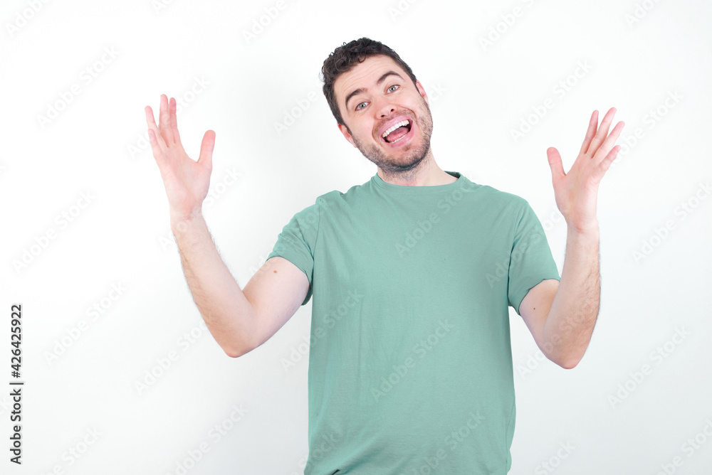 Cheerful young handsome caucasian man wearing green t-shirt against white background making a welcome gesture raising arms over head. Cheerful young handsome caucasian man wearing green t-shirt against white background making a welcome gesture raising arms over head.