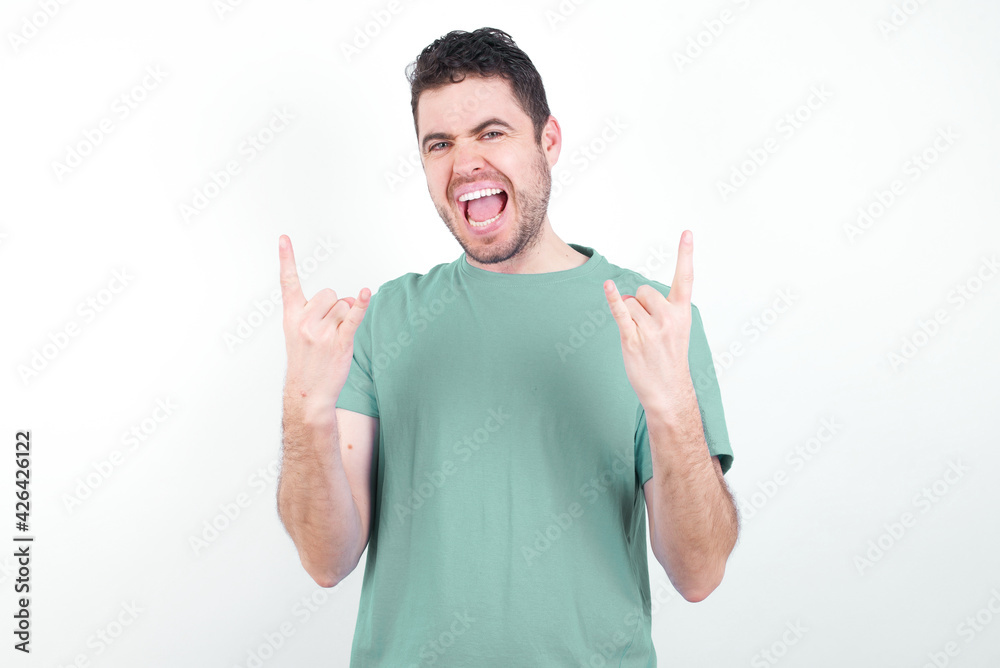 Born to rock this world. Joyful young handsome caucasian man wearing green t-shirt against white background screaming out loud and showing with raised arms horns or rock gesture. Born to rock this world. Joyful young handsome caucasian man wearing green t-shirt against white background screaming out loud and showing with raised arms horns or rock gesture.