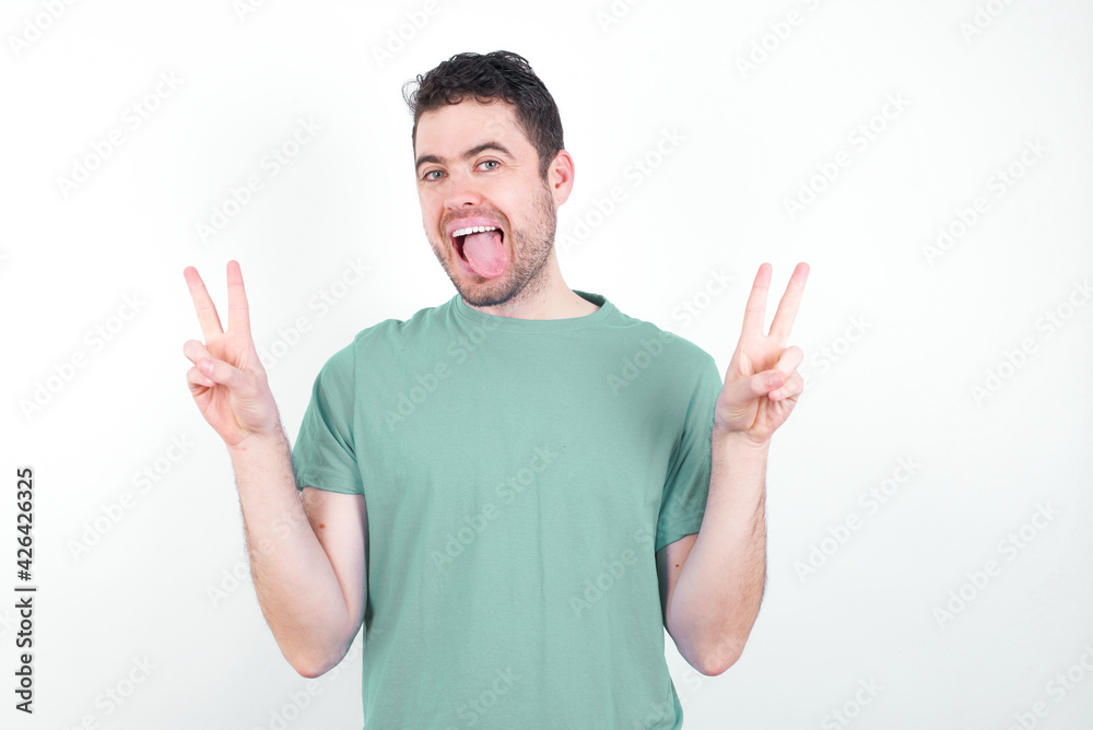 Fototapeta premium young handsome caucasian man wearing green t-shirt against white background with optimistic smile, showing peace or victory gesture with both hands, looking friendly. V sign.