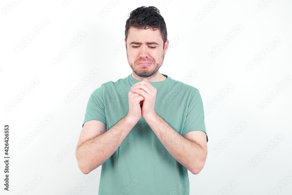 Sad young handsome caucasian man wearing green t-shirt against white background feeling upset while spending time at home alone staring at camera with unhappy or regretful look. Sad young handsome caucasian man wearing green t-shirt against white background feeling upset while spending time at home alone staring at camera with unhappy or regretful look.