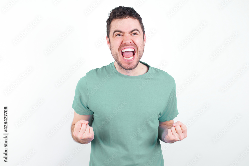 young handsome caucasian man wearing green t-shirt against white background raising fists up screaming with joy being happy to achieve goals. young handsome caucasian man wearing green t-shirt against white background raising fists up screaming with joy being happy to achieve goals.