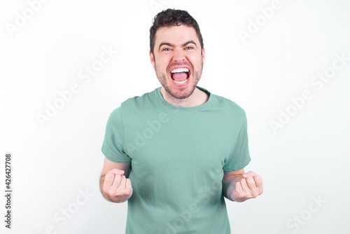 young handsome caucasian man wearing green t-shirt against white background raising fists up screaming with joy being happy to achieve goals.