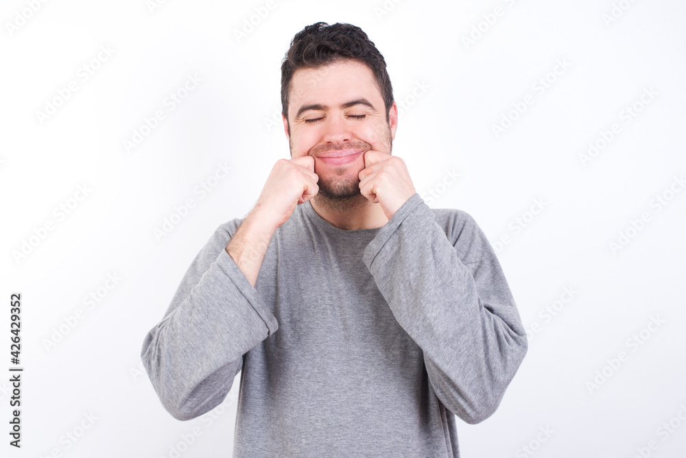 Pleased young handsome caucasian bearded man wearing pyjama over white wall with closed eyes keeps hands near cheeks and smiles tenderly imagines something very pleasant Pleased young handsome caucasian bearded man wearing pyjama over white wall with closed eyes keeps hands near cheeks and smiles tenderly imagines something very pleasant