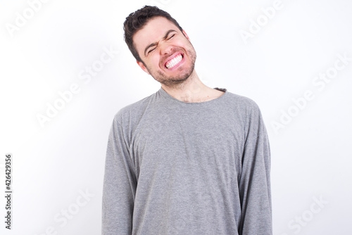 Positive young handsome caucasian bearded man wearing pyjama over white wall with overjoyed expression closes eyes and laughs shows white perfect teeth