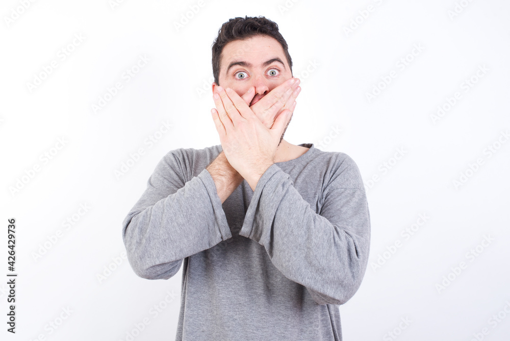 Stunned young handsome caucasian bearded man wearing pyjama over white wall covers both hands on mouth, afraids of something astonishing Stunned young handsome caucasian bearded man wearing pyjama over white wall covers both hands on mouth, afraids of something astonishing