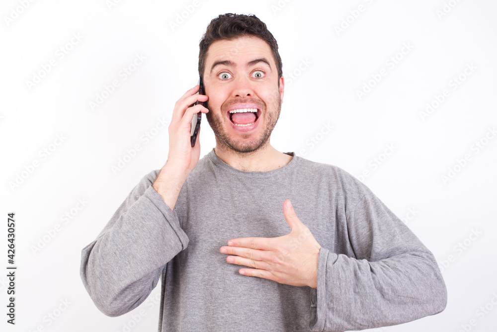 Smiling young handsome caucasian bearded man wearing pyjama over white wall talks via cellphone, enjoys pleasant great conversation. People, technology, communication concept