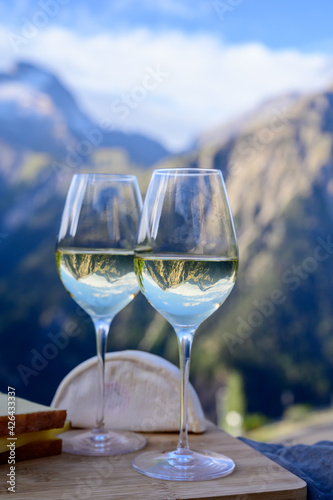 Tasty cheese and wine from Savoy region in France, beaufort, abondance, emmental, tomme and reblochon de savoie cheeses and glass of white wine served outdoor with Alpine mountains peaks on background