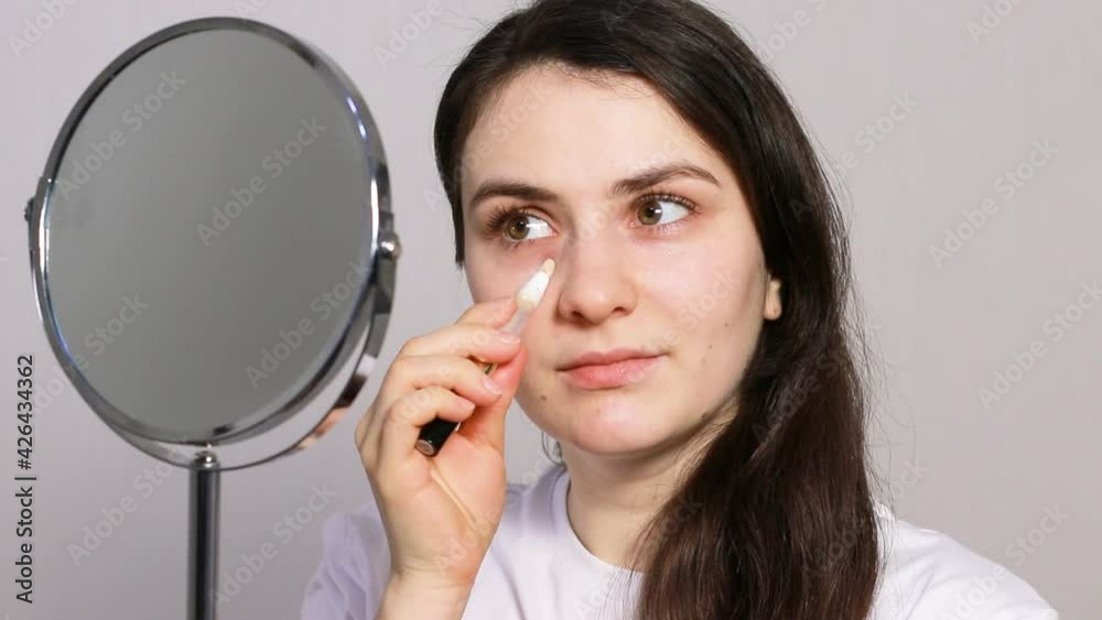Young beautiful woman applying concealer under the eyes while sitting in front of the mirror. Decorative cosmetics for the skin, make-up and concealment of skin imperfections, bruises and puffiness Young beautiful woman applying concealer under the eyes while sitting in front of the mirror. Decorative cosmetics for the skin, make-up and concealment of skin imperfections, bruises and puffiness