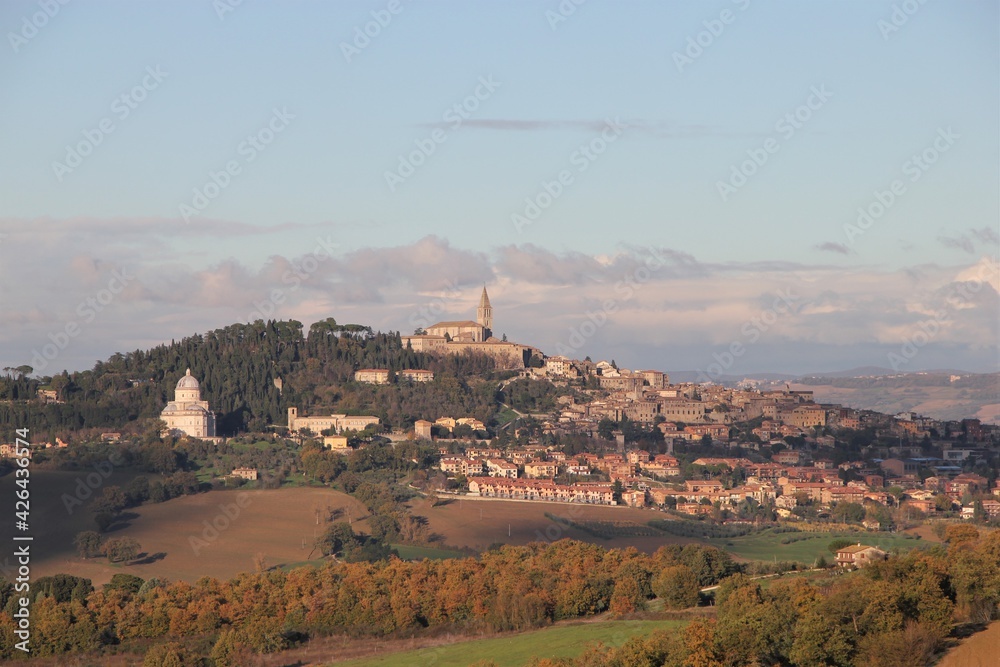 The iconic Umbrian hill town of Todi seen from the West dominated by Consolazione basilica and San Fortunato belltower, surrounded by autumn colored trees, torn clouds in the distance
