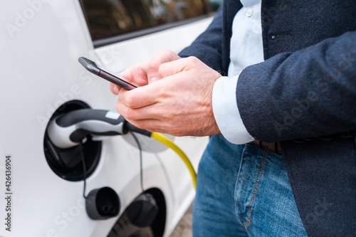 Close up a man in the suit standing near the charging electric car and using his smartphone for paying 
