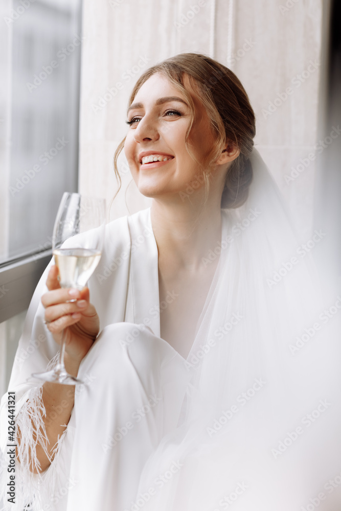 Bride s morning. Bride drinking champagne in the peignoir. young woman is sitting on a large window in a hotel room in bathroom. Beautiful girl in white wedding robe. wedding day Bride s morning. Bride drinking champagne in the peignoir. young woman is sitting on a large window in a hotel room in bathroom. Beautiful girl in white wedding robe. wedding day
