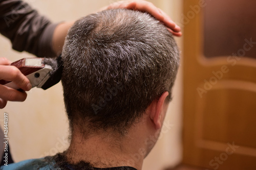 A man is trimmed with a hair clipper and sits with his back to the camera. Perhaps at home in a lockdown