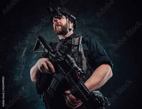 Soldier with helmet and vest stays holding a rifle in dark background