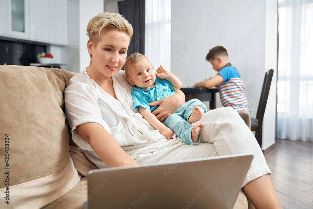 Pleasant woman with blond hair working on laptop and carrying toddler on hands. Blur background of teenage boy sitting on table and doing homework.