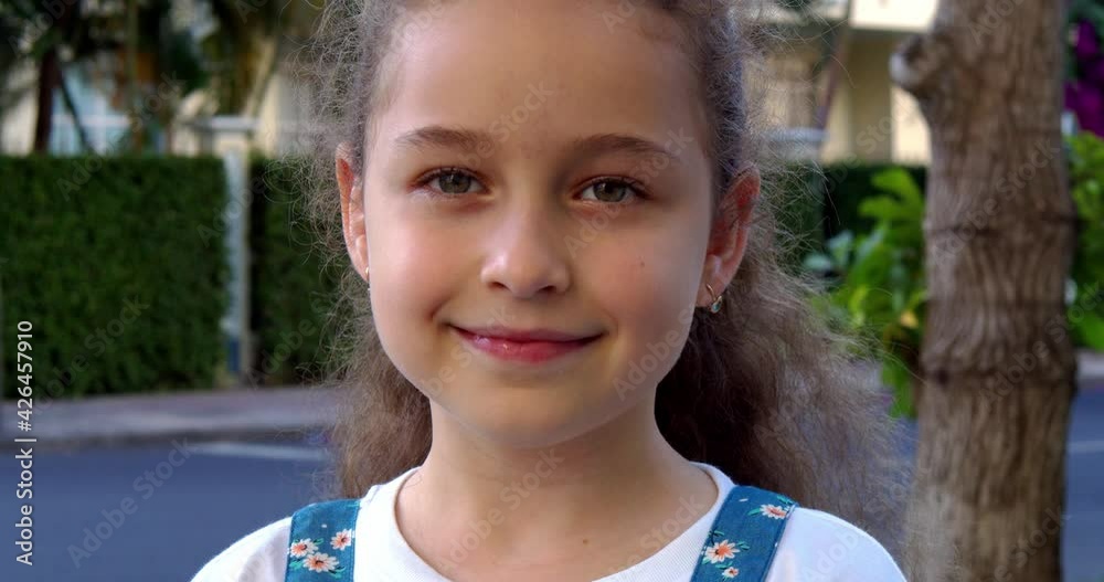 Portrait cute little child girl looking at camera standing on street in city on summer day. Young happy thinking curiosity child looking at camera closeup outdoors.