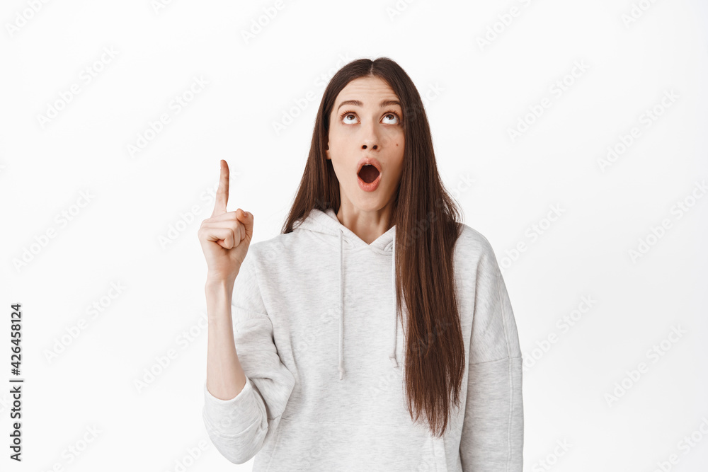 Wow, thrilled girl showing awesome promo deal, pointing and looking up at something cool, gasping amazed, checking out promotion, standing over white background Wow, thrilled girl showing awesome promo deal, pointing and looking up at something cool, gasping amazed, checking out promotion, standing over white background