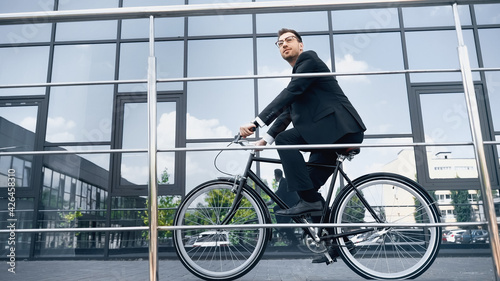 full length of businessman in suit riding bicycle near building with glass facade.