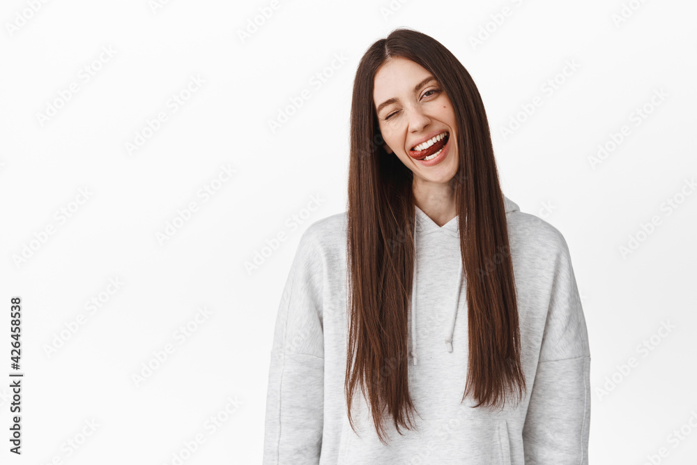 Joyful teenage girl student with long brown hair, tilt head, wink at you and shows tongue positive, express happiness and joy, standing over white background Joyful teenage girl student with long brown hair, tilt head, wink at you and shows tongue positive, express happiness and joy, standing over white background