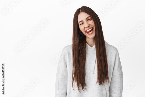 Joyful teenage girl student with long brown hair, tilt head, wink at you and shows tongue positive, express happiness and joy, standing over white background © Cookie Studio