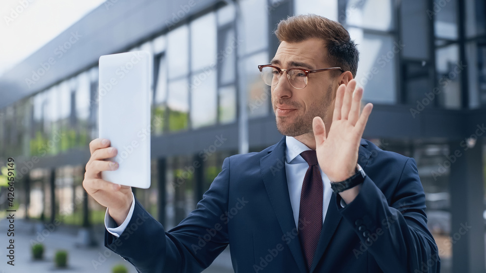 businessman in glasses waving hand while having video call and holding digital tablet outside. businessman in glasses waving hand while having video call and holding digital tablet outside.