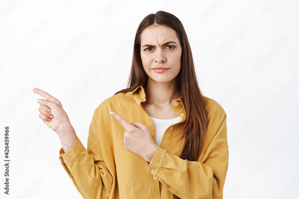 Skeptical and doubtful woman frowning, pointing fingers aside at something strange, hesitating, being unsure about product, standing against white background Skeptical and doubtful woman frowning, pointing fingers aside at something strange, hesitating, being unsure about product, standing against white background