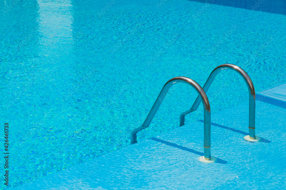 Swimming pool with stair and blue water. Grab bars ladder in the blue swimming pool. Ripple wave in pool for background and abstract. Summer relax concept. Texture of water surface. Space for text.