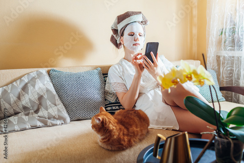 Woman with facial sheet mask applied relaxing at home after bath sitting on couch with cat using phone. Skincare routine