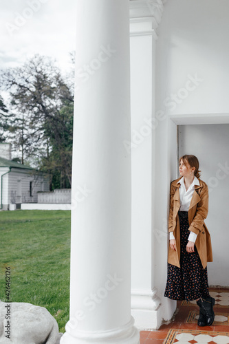  Stylish young girl stands near a column against a white wall and looks up the hill