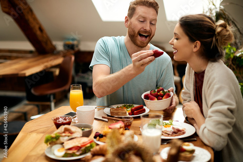 Happy man feeding his girlfriend with strawberries during breakfast at dining table.