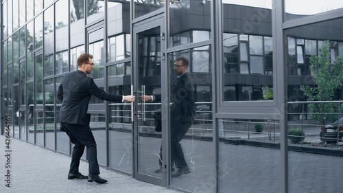 full length of businessman in suit opening door in building with glass facade.