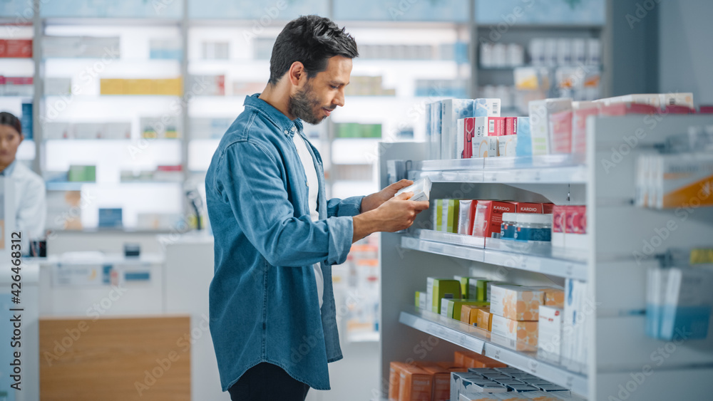 Pharmacy Drugstore: Portrait of Handsome Young Latin Man Searching to Purchase Best Medicine, Chooses between Two Packages of Drugs, Vitamins. Shelves full of Health Care, Wellness, Sport Supplements Pharmacy Drugstore: Portrait of Handsome Young Latin Man Searching to Purchase Best Medicine, Chooses between Two Packages of Drugs, Vitamins. Shelves full of Health Care, Wellness, Sport Supplements