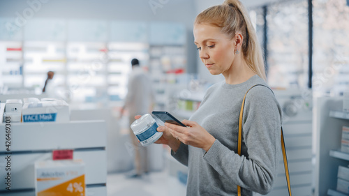 Pharmacy Drugstore: Portrait of a Beautiful Caucasian Woman Using Smartphone Search to Best Cream, Checks Jar for Eco Firendly Clean Ingredients and Proceeds to Checkout Counter With Happy Purchase photo