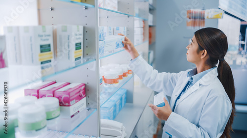 Pharmacy Drugstore: Portrait of Beautiful Diligent Asian Pharmacist Wearing White Coat Arranges Medicine Packages, Drug Boxes, Vitamin and Supplement Pills on a Shelf Behind the Counter