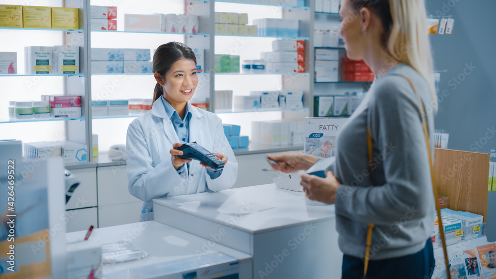 Pharmacy Drugstore Checkout Cashier Counter: Professional Asian Female Pharmacist Recommends Medicine in a Package, Caucasian Female Customer Pays Using Contactless Payment Terminal and Credit Card
