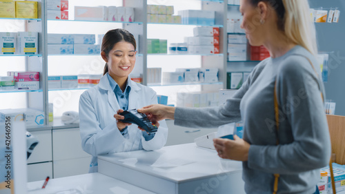 Pharmacy Drugstore Checkout Cashier Counter: Professional Asian Female Pharmacist Recommends Medicine in a Package, Caucasian Female Customer Pays Using Contactless Payment Terminal and Credit Card