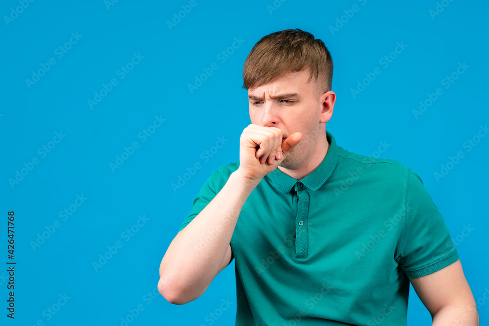 A young patient, a man coughing, holds his hands in a fist to his face. The guy suffers from bronchitis or pneumonia, blue background. Cold, Chinese coronavirus virus, flu, disease concept. A young patient, a man coughing, holds his hands in a fist to his face. The guy suffers from bronchitis or pneumonia, blue background. Cold, Chinese coronavirus virus, flu, disease concept.