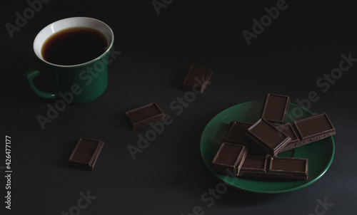 A cup of strong hot coffee with pieces of dark chocolate on a saucer on a dark background. Green teacup and saucer. Atmospheric horizontal photo, top view. 