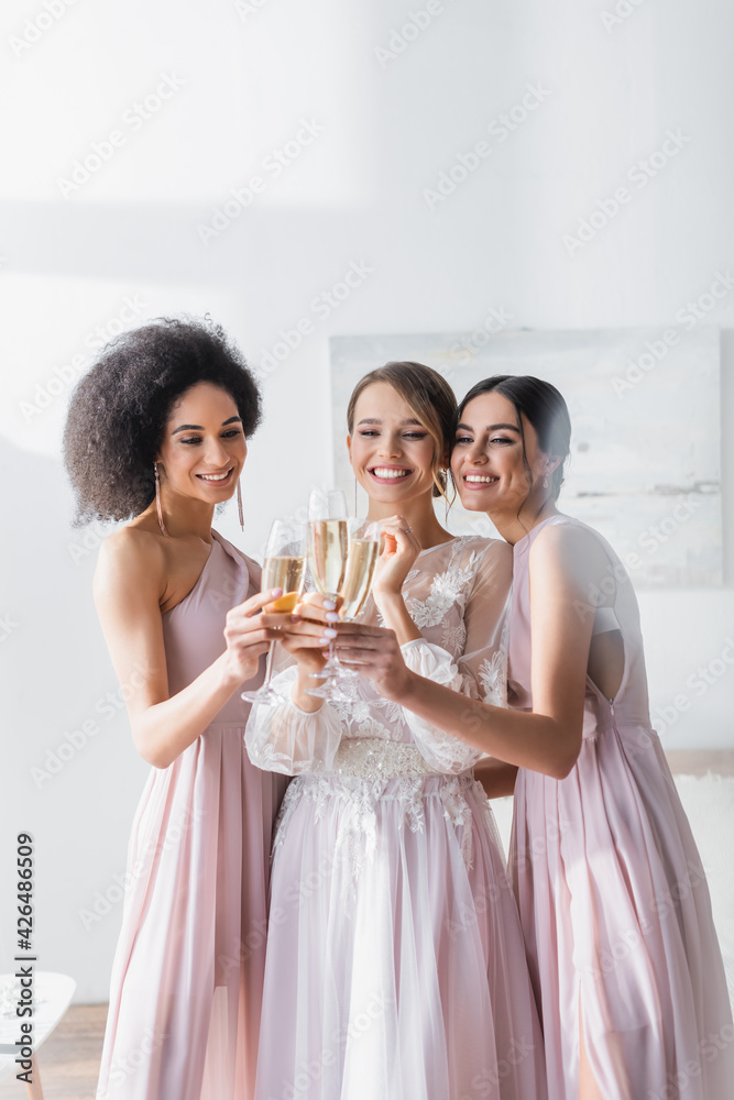 joyful interracial bridesmaids clinking champagne glasses with bride in bedroom. joyful interracial bridesmaids clinking champagne glasses with bride in bedroom.