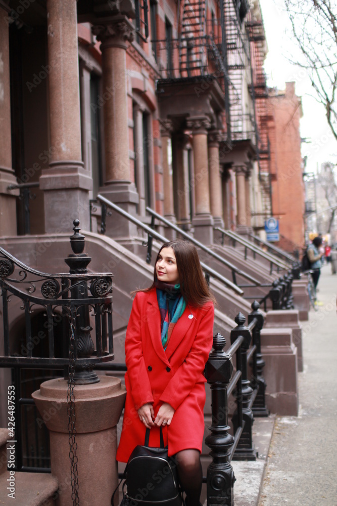a caucasian woman with dark hair and wearing a red coat stands outside in the city, on a sparsely populated street. a stylish woman traveling alone or with a friend and enjoying life, modern